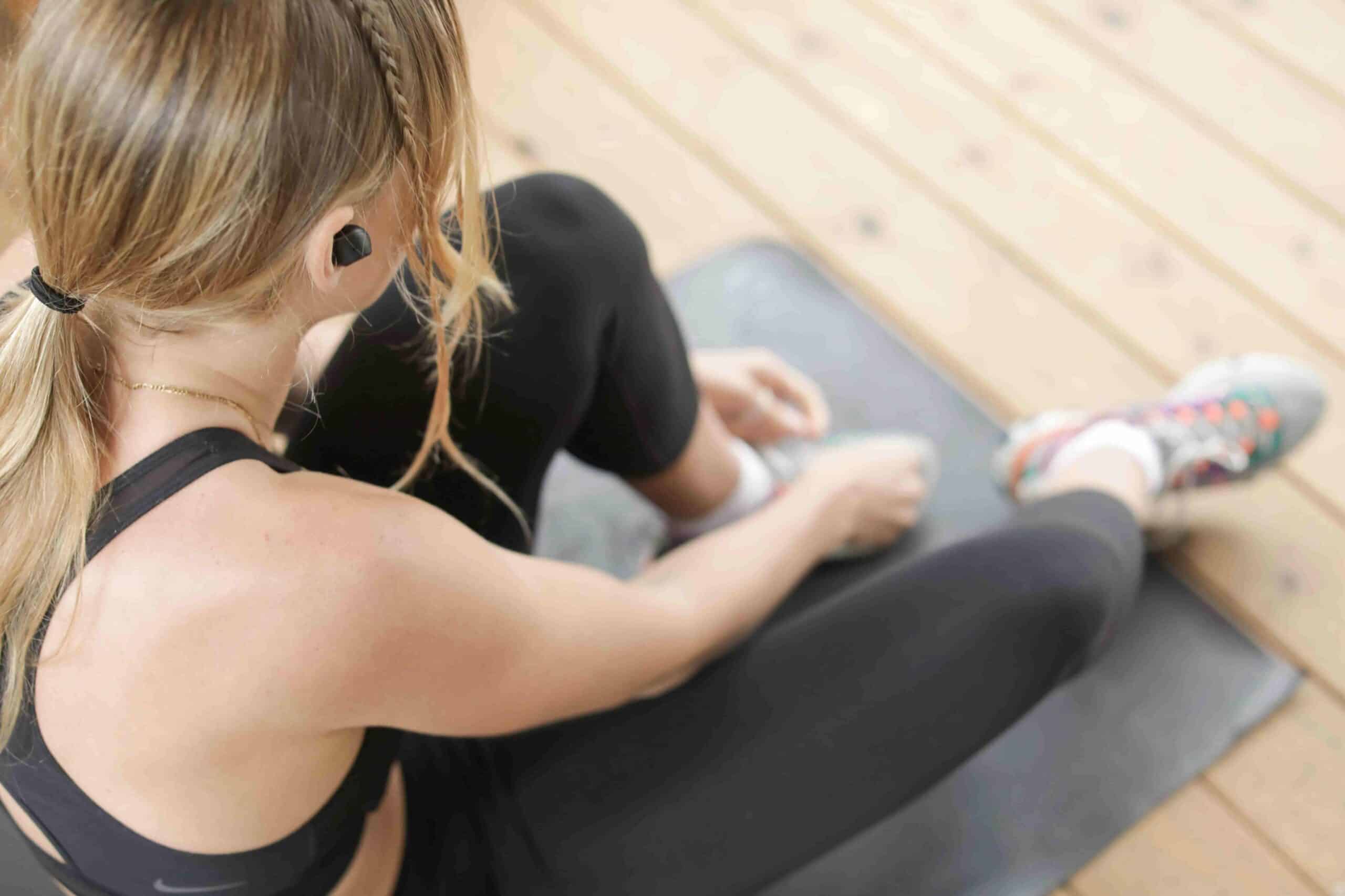 A woman in athletic wear sits on a yoga mat outdoors, tying her shoelaces, with wireless earbuds in her ears.