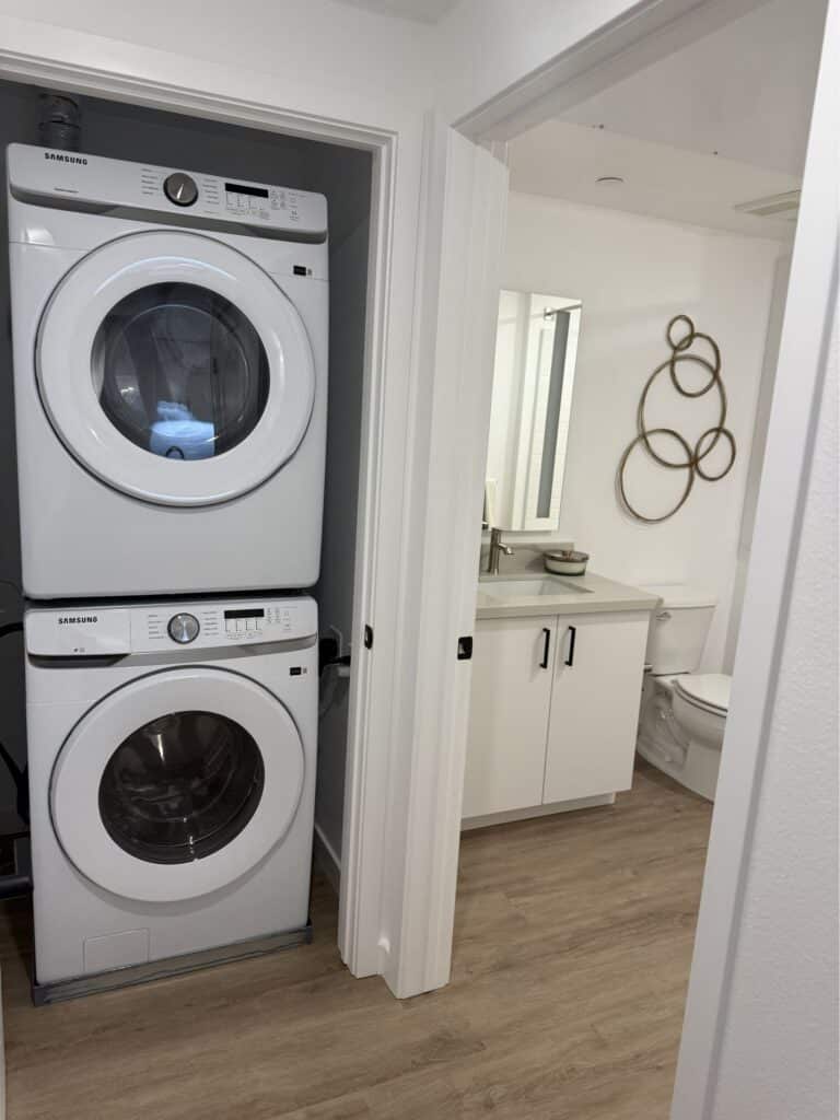 A stacked washer and dryer are in a laundry closet next to a bathroom with a white vanity, toilet, and decorative wall art. The floor is light wood.