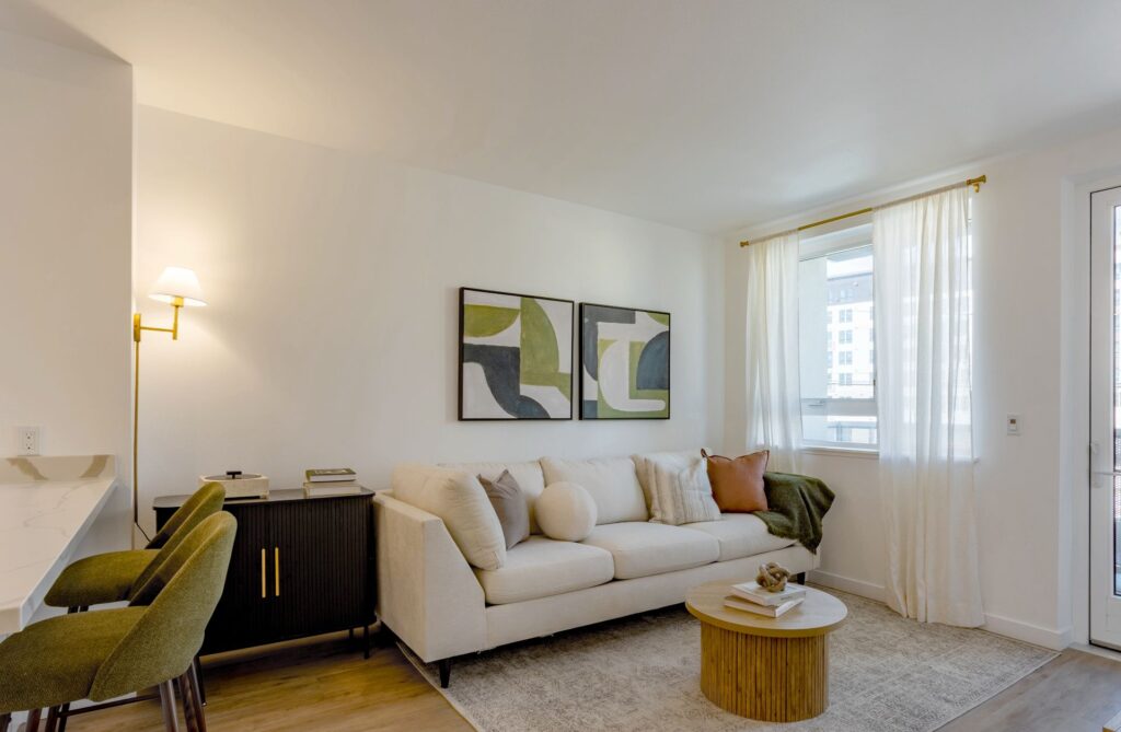 A modern living room with a beige sofa, round coffee table, two abstract paintings on the wall, green chairs, and a window with sheer curtains.