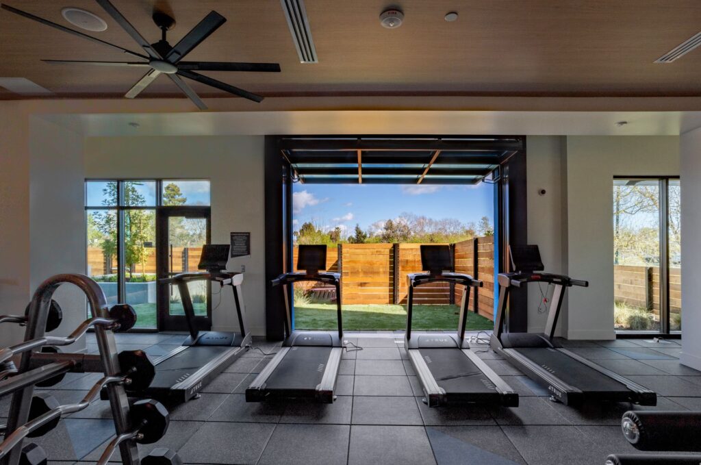Four treadmills are positioned in front of a large open window in a gym, with weights visible on the left and a wooden fence and trees outside.
