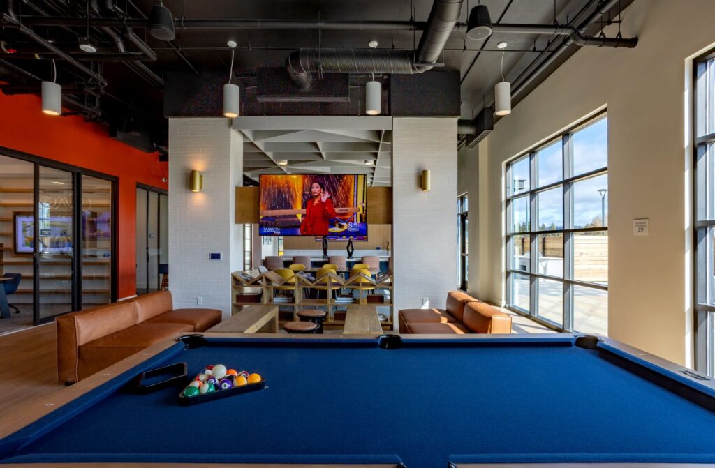 A modern game room with a blue pool table in the foreground, brown sofas, a TV mounted on the wall, and large windows letting in natural light.
