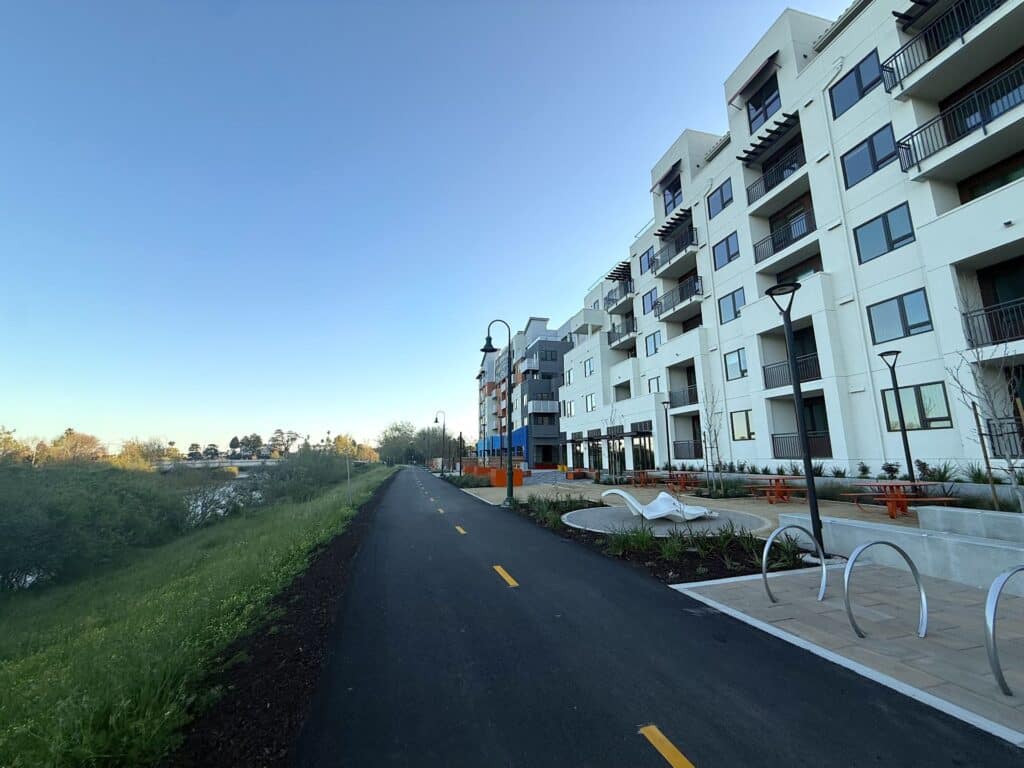 A paved bike and pedestrian path runs alongside a modern multi-story apartment building with balconies and outdoor seating areas.